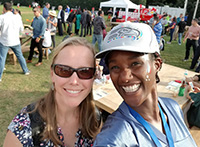 Two women, one wearing sunglasses, the other wearing a ballcap, stand together; crowd of people in background