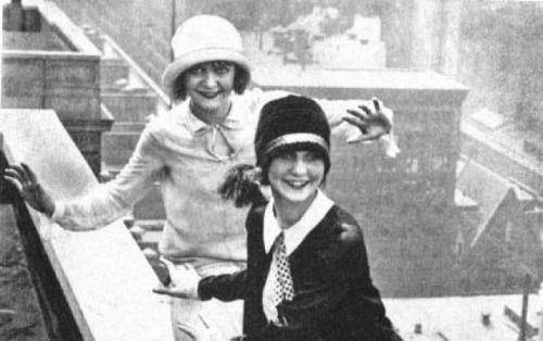 B/W photo of two1920s flappers dancing on a skyscraper ledge