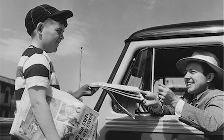 Black & white photo of newsboy selling tabloid newspaper to man in car wearing white fedora