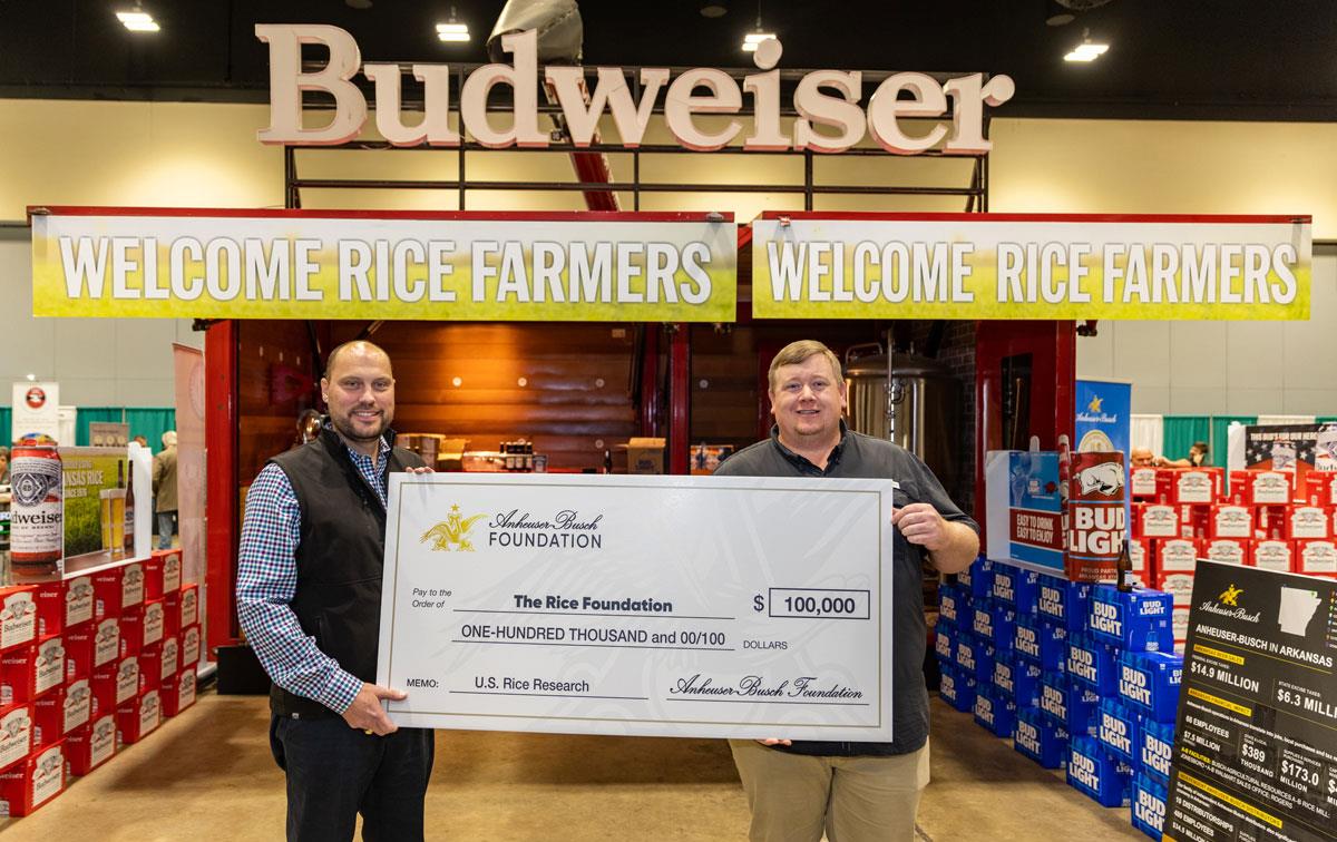 AB-Donation-to-Rice-Foundation,-Brian-Ottis-&-Bill-Jones hold big check while standing in front of the Budweiser Bar with signs reading "Welcome Rice Farmers"