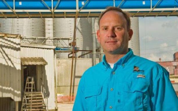 White man wearing blue shirt stands in front of rice mill