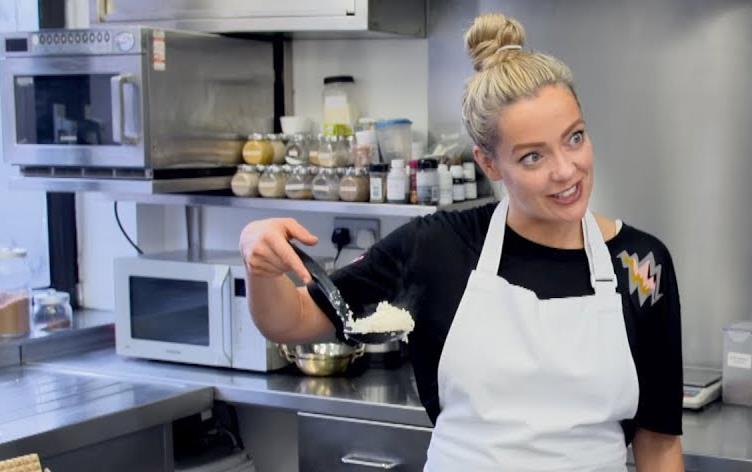 White woman wearing white apron, standing in a kitchen, holds spoonful of white rice