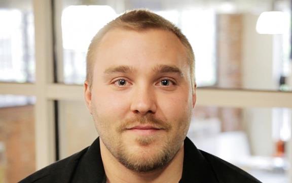 Headshot of white male with close cropped hair and a goatee wearing an open collar black shirt with company insignia