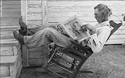 Vintage photo of old man wearing overalls & reading newspaper, rocking back in rocking chair with his feet on steps