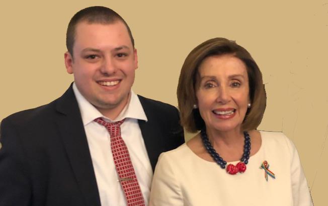 Intern Grant Long, wearing suit and tie, stands next to Speaker of the House Nancy Pelosi, wearing white dress, carrying small brown bag