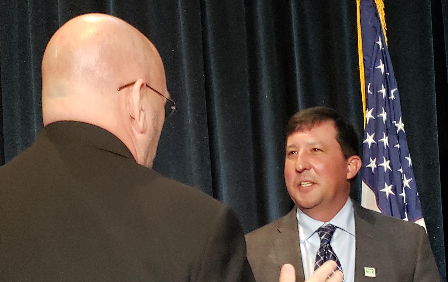 Two white men wearing business suits engaged in animated conversation, American flag in background