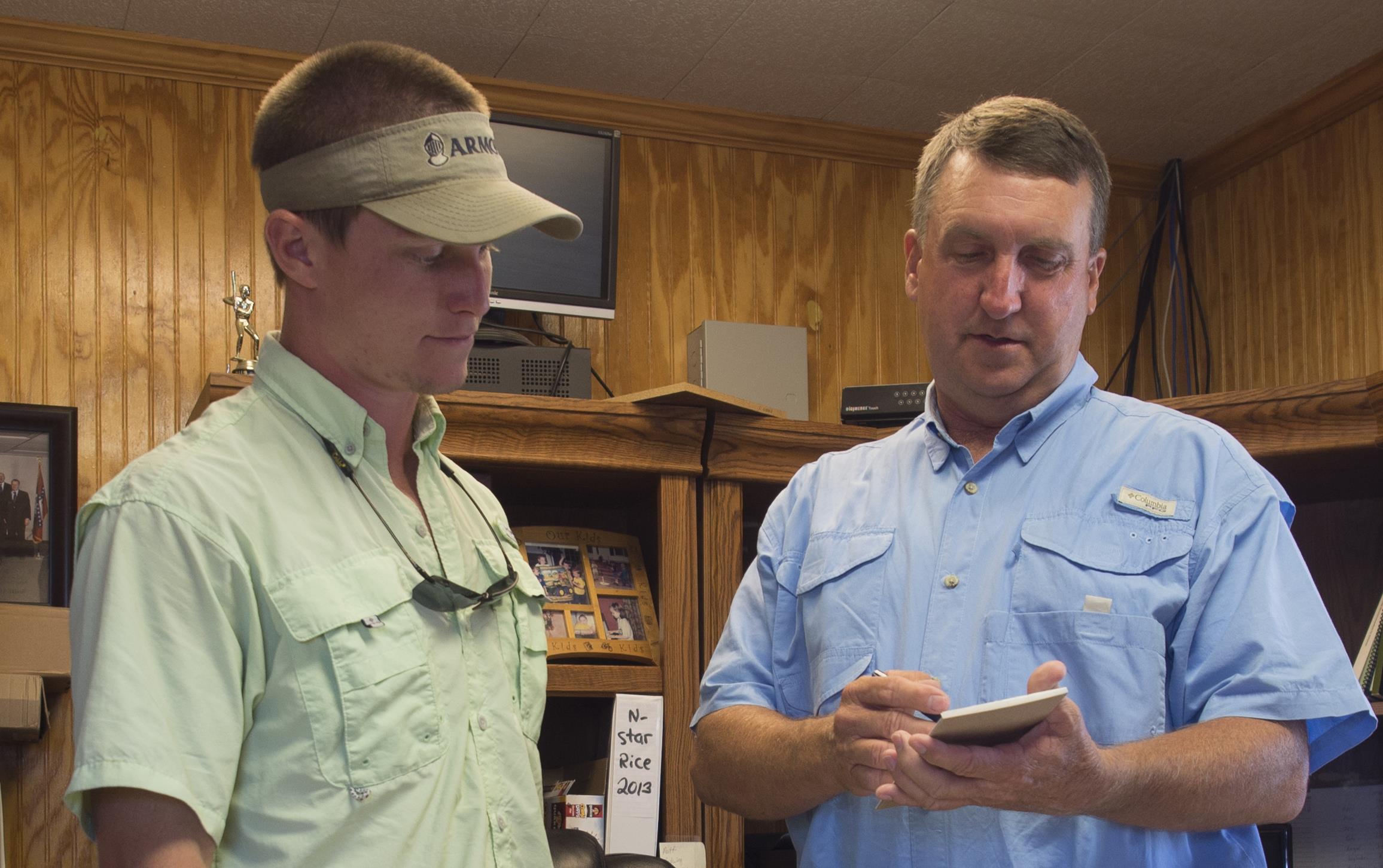 Two men dressed in casual shirts and pants stand next to desk in wood paneled office, checking details on a pad of paper