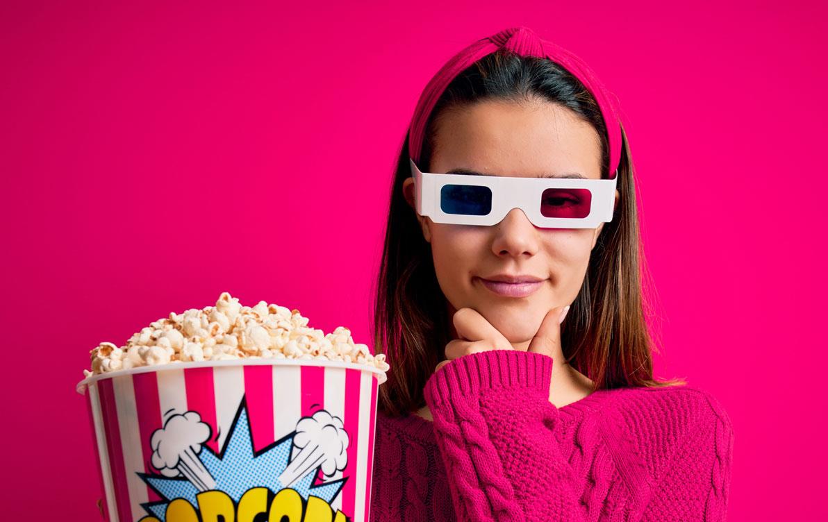Girl wearing fuchsia sweater & 3D glasses holds large popcorn bucket, fuchsia background