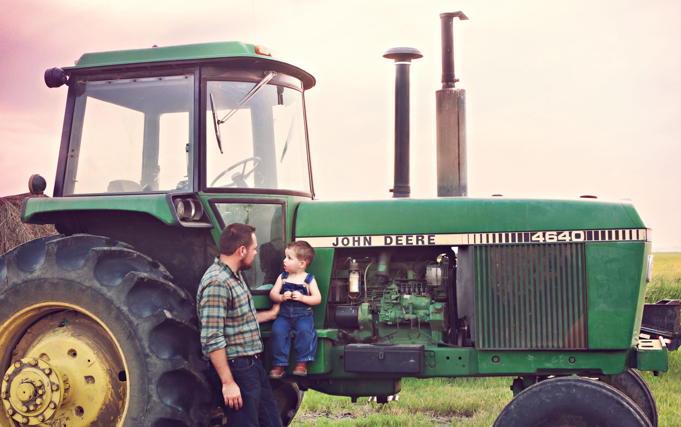 Father holds son wearing overalls up against vintage John Deere tractor