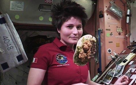 Female astronaut Samantha Cristoforetti wearing maroon shirt, cooks in zero gravity with food floating in space