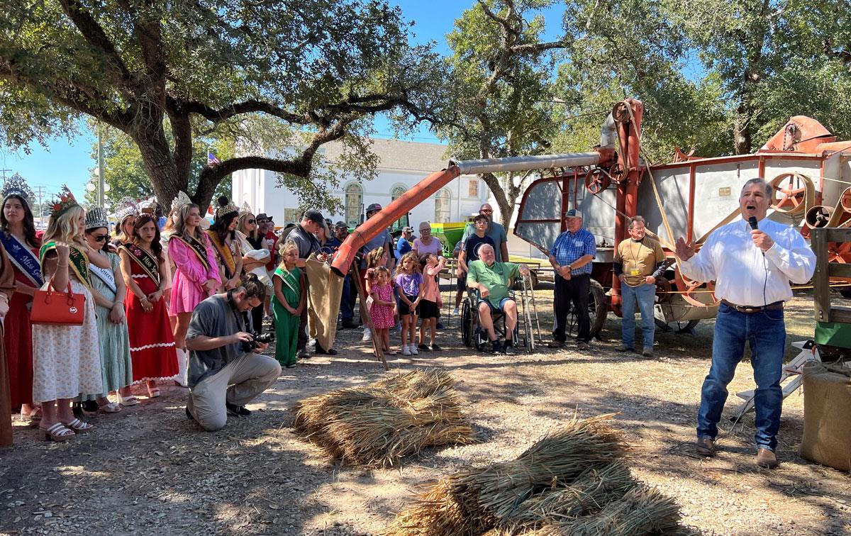 Crowd gathers to watch old fashioned rice threshing demo, Ronnie-Levy-with-mic, stacks of threshed stalks in foreground