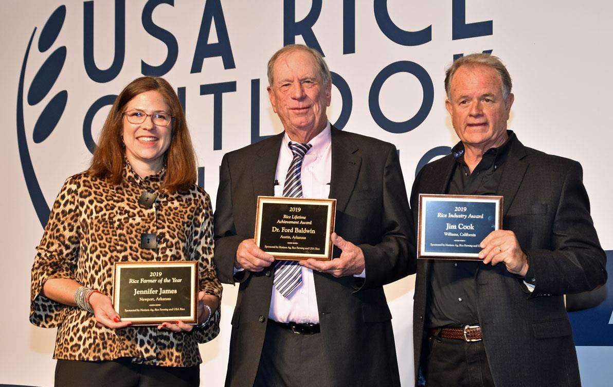 A woman and two men stand together holding awards plaques