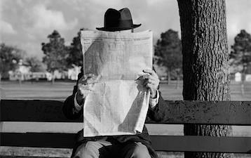 Black & white photo of man wearing a top hat, sitting on bench reading newspaper