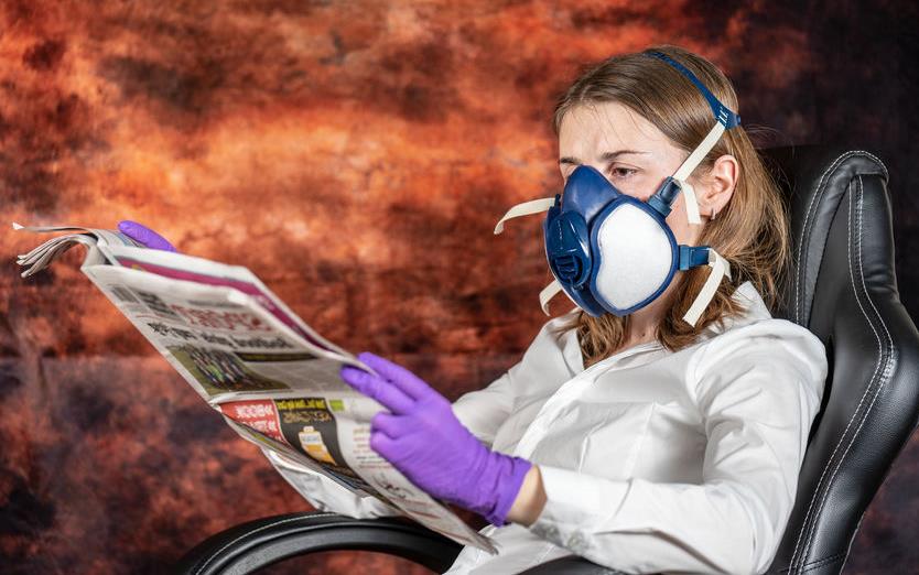 Woman wearing gas mask and purple gloves sits in chair reading a newspaper