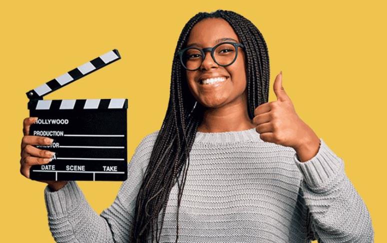 Young, black female student holds movie clapboard and does thumbs up, yellow bkgd