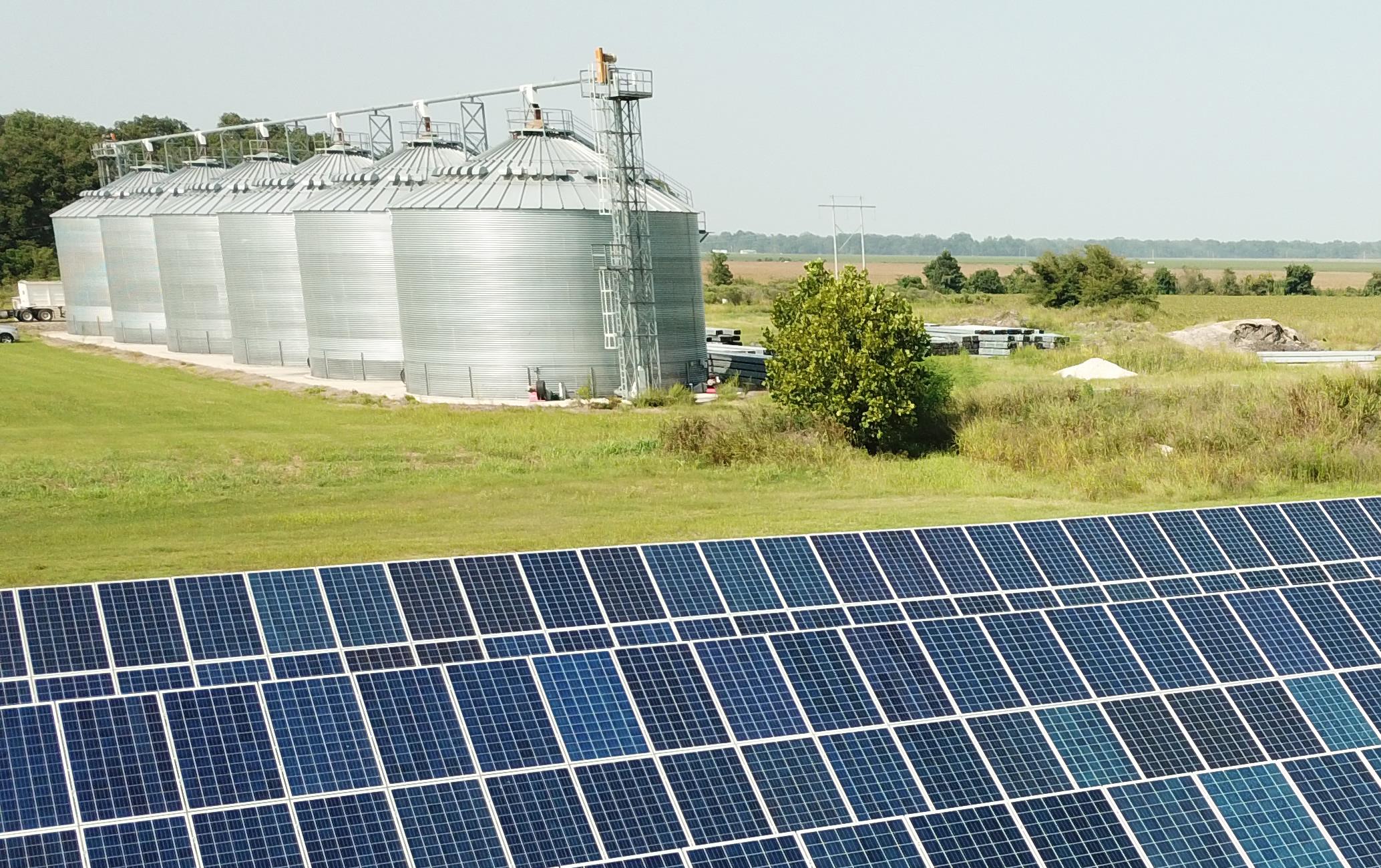 Rows of blue solar panels in a green field with grain bins in the background