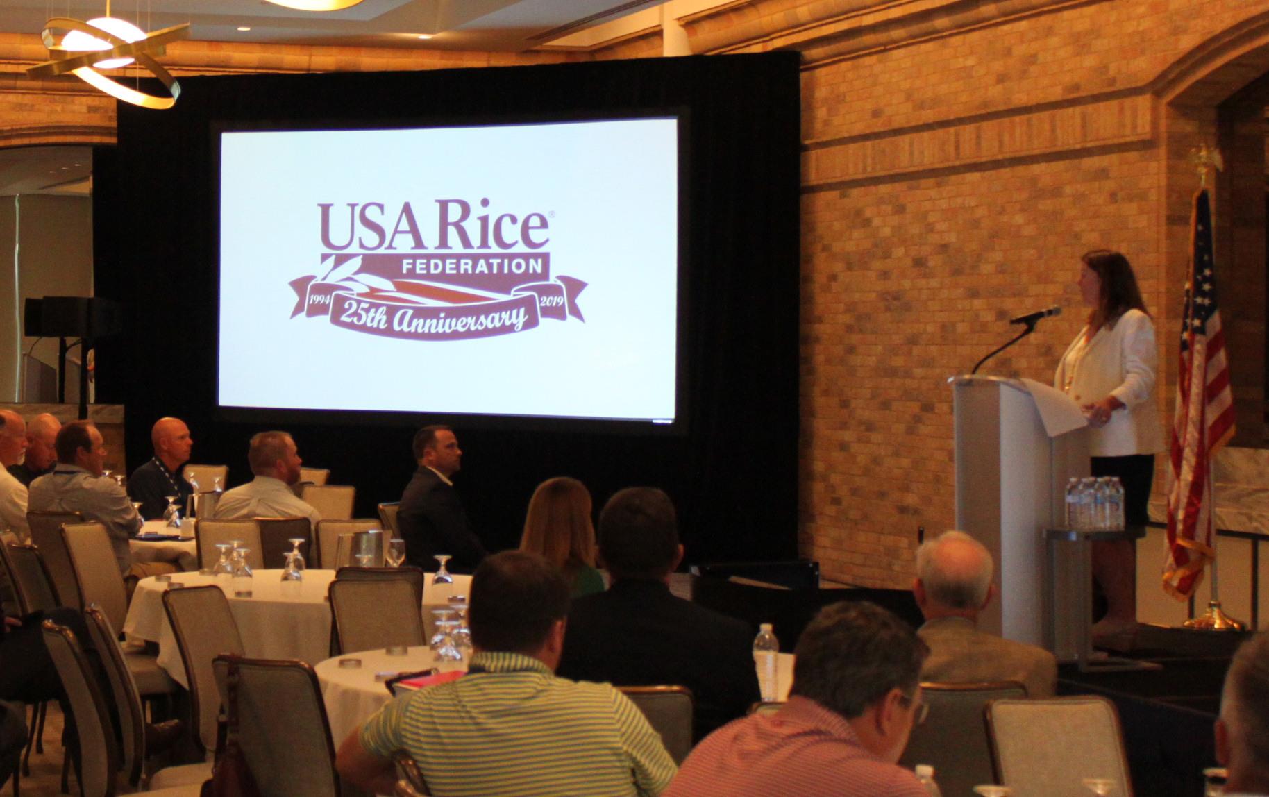Crowded meeting room, woman standing at podium next to projected slides that reads: "USA Rice Federation 25th Anniversary"