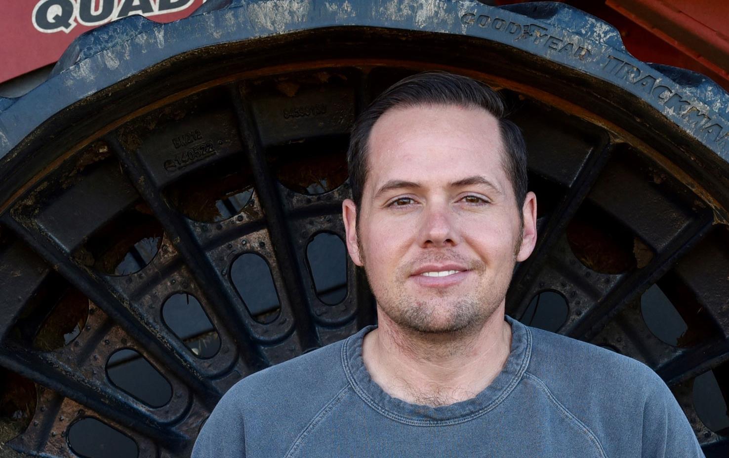 Young white male wearing gray sweatshirt standing in wheel well of Case combine