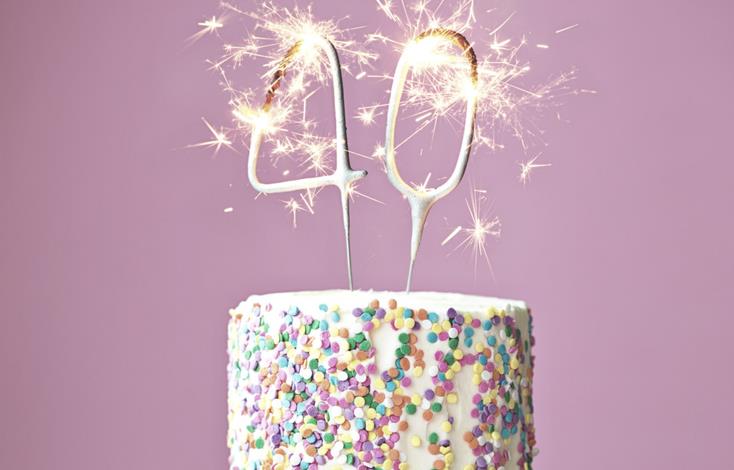 White cake covered with sprinkles & sparklers in the shape of "40" sits on white cake stand, mauve background