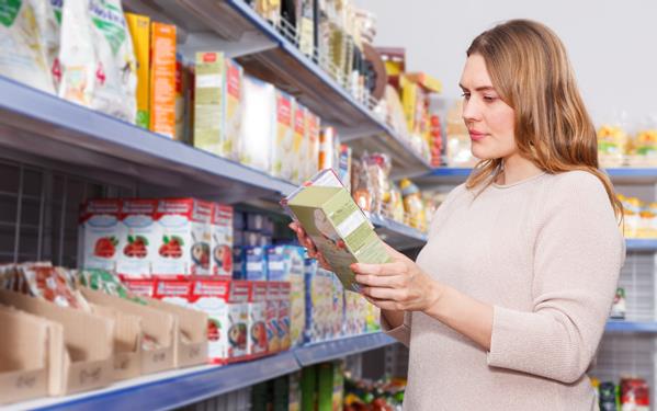 Woman in grocery store full of stocked shelves looking at back of box ingredient list