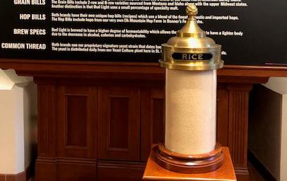 Large glass canister with brass top and label filled with white rice sits onto a pedestal in front of poster describing Anheuser-Busch brewing process