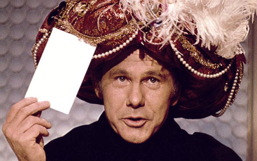 Johnny Carson as Carnac the Magnificent wearing jeweled turban, holding envelope in one hand