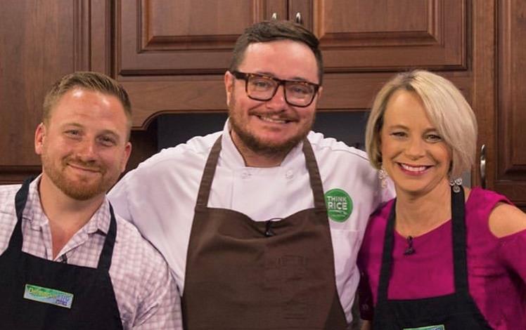 Chef Hari Cameron wearing Think Rice chef coat and brown apron stands with his arms around a man and a woman wearing black aprons