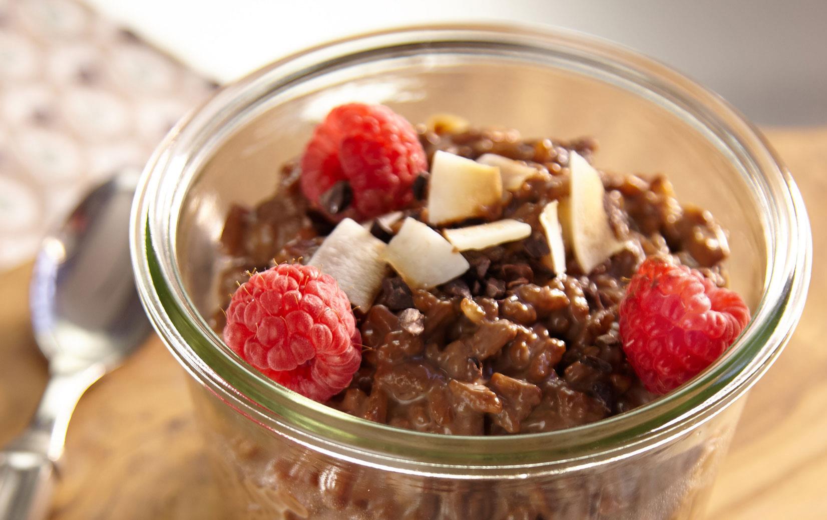 Chocolate-Espresso-Porridge with raspberries and slivered almonds in a glass dish sitting on a small wooden cutting board on top of a print napkin