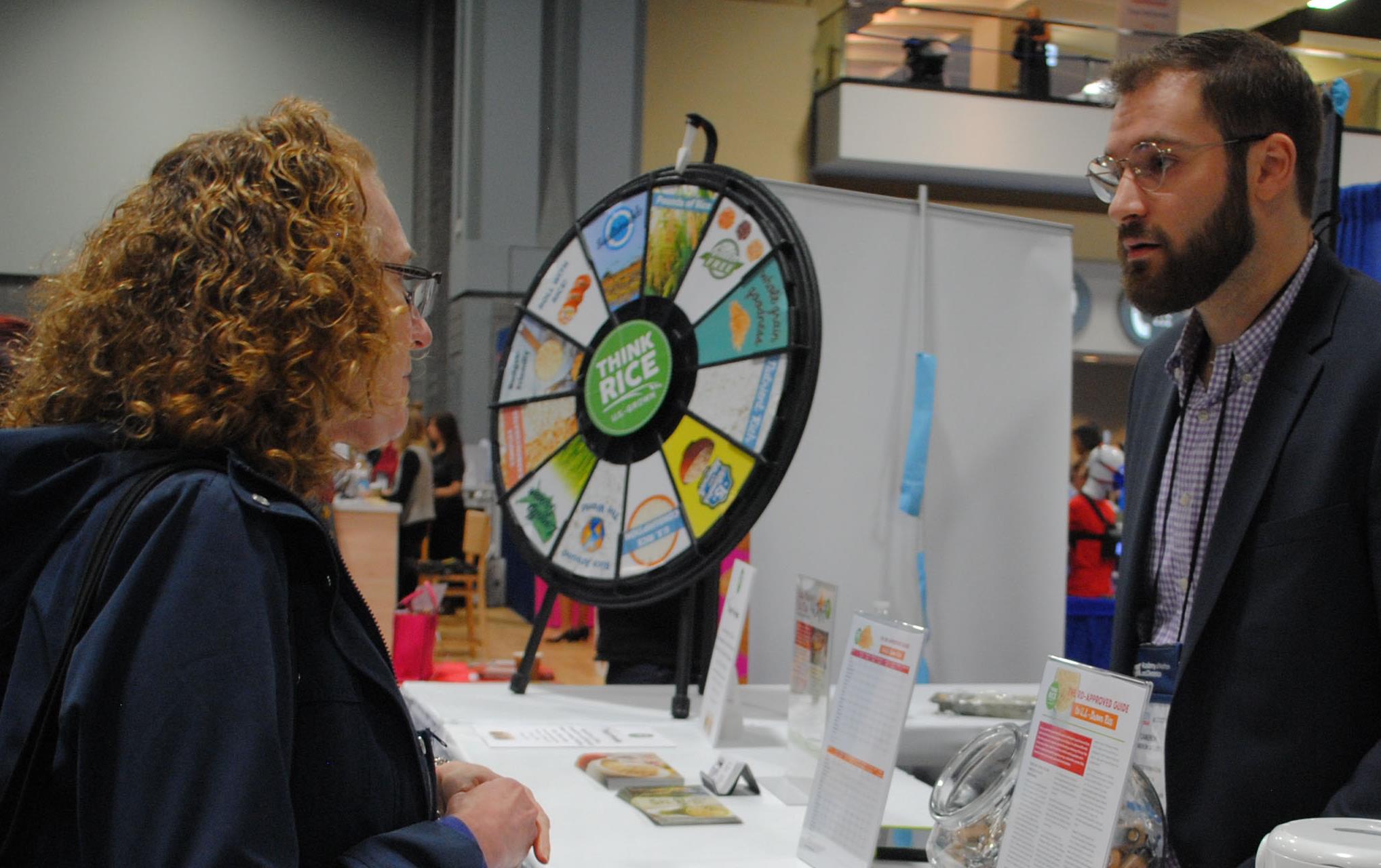 Man with glasses wearing blue blazer talks with red-headed woman carrying multicolor bag, at trade show booth with table display on different rice types and Think Rice trivia wheel