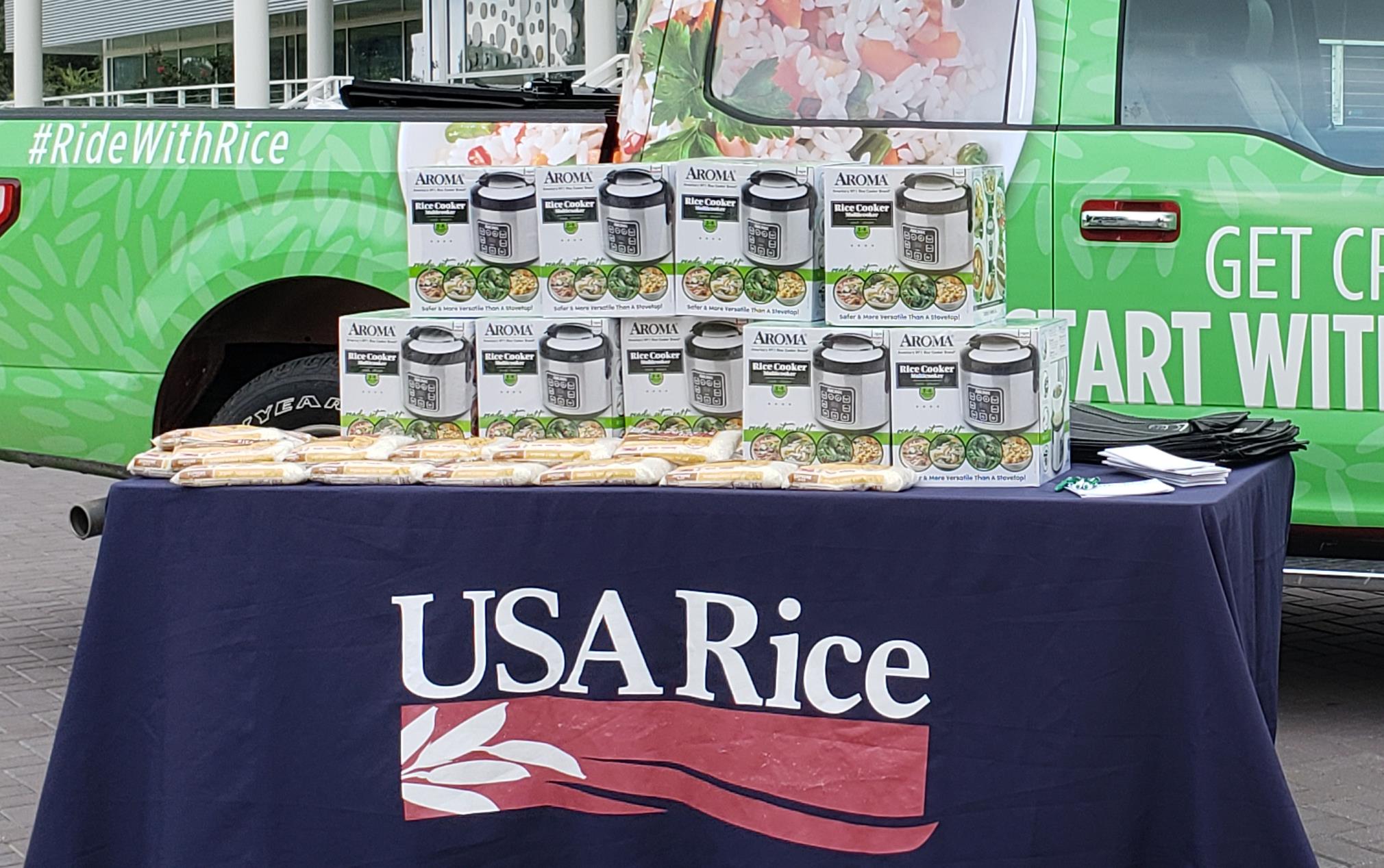 Green pickup truck with designs & words "Get Creative, Start With Rice," sits behind table with USA Rice banner and stacks of boxes on top