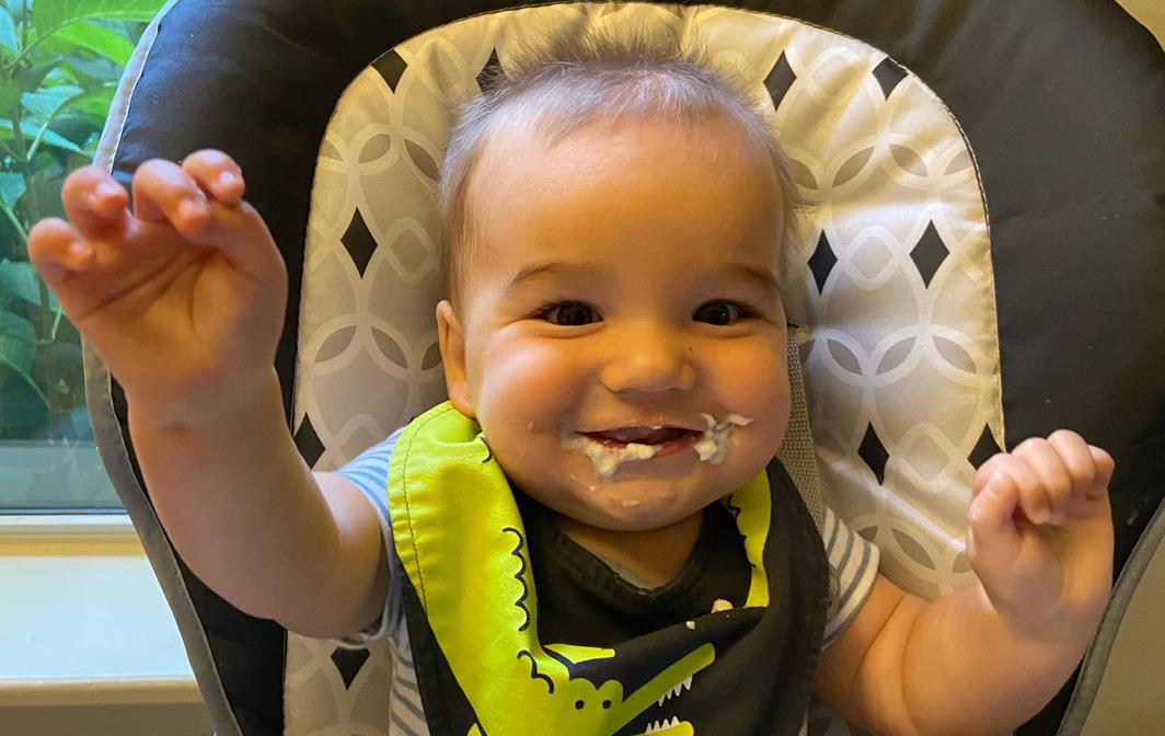 Smiling baby with rice on his face reaches his hands up in the air, wearing colorful bib with green dragon