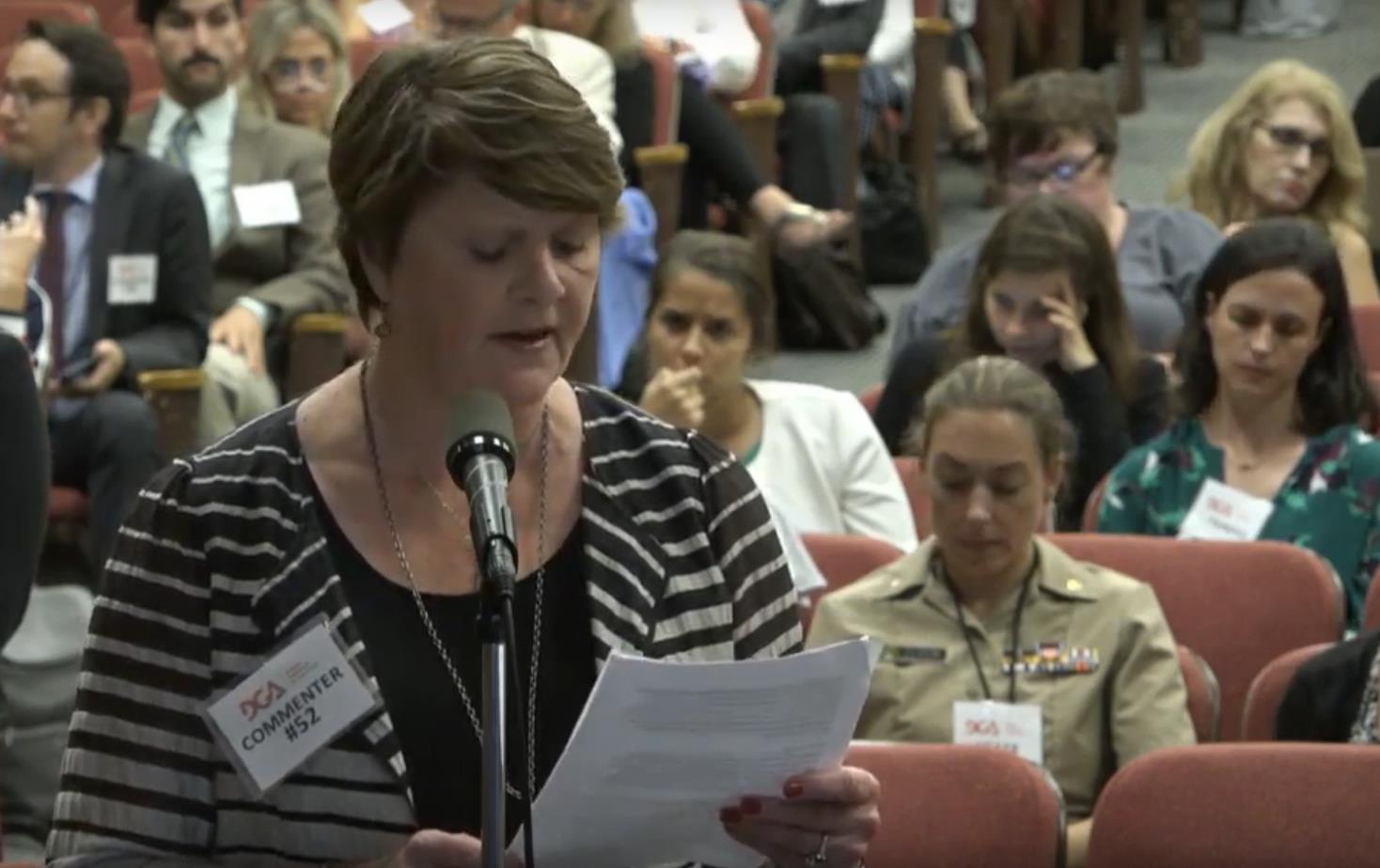 White woman with short brown hair, wearing a black and white striped shirt, stands in front of a microphone reading from a paper in her hand, large group seated behind her