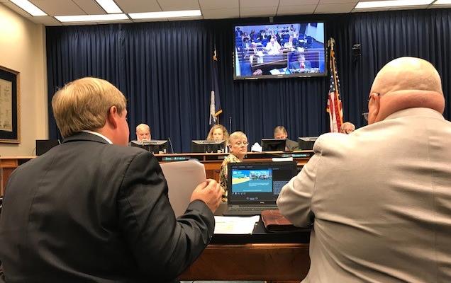 Two men in suits with their backs to the camera sit at a desk in front of a panel of people and a bank of video screens