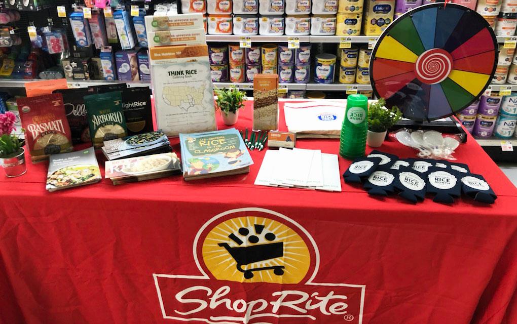 ShopRite demo table filled with bags of rice, coloring books, cups, brouchures, and game wheel