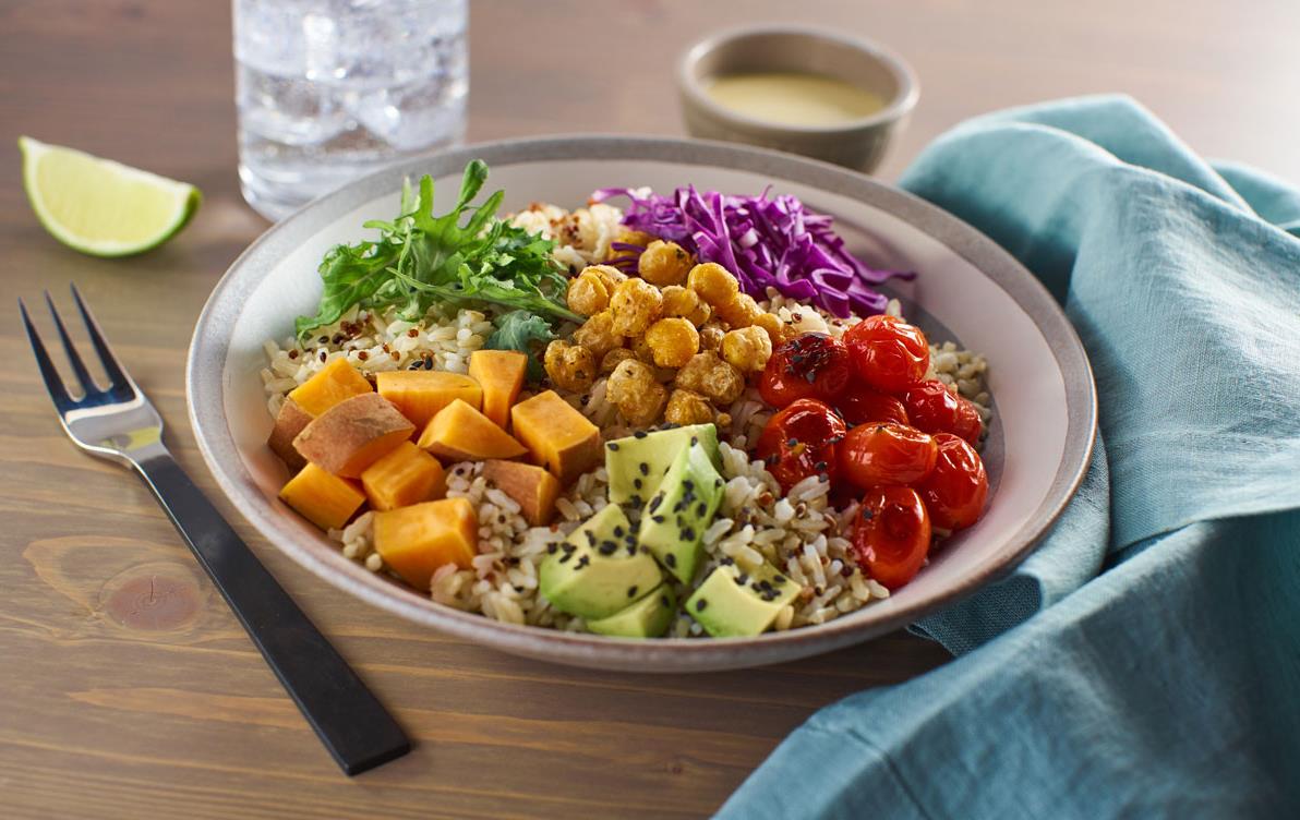 Bowl filled with rice and colorful veggies sits on table with blue napkin, silver fork, glass of water