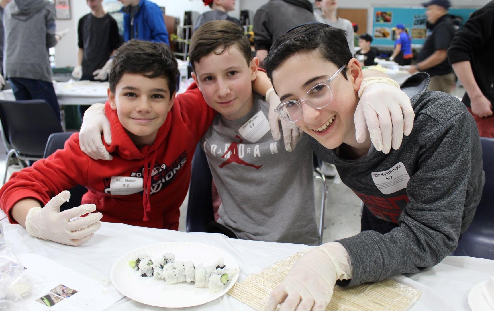 Three boys with their arms around each others shoulders sit in front of plate of homemade sushi