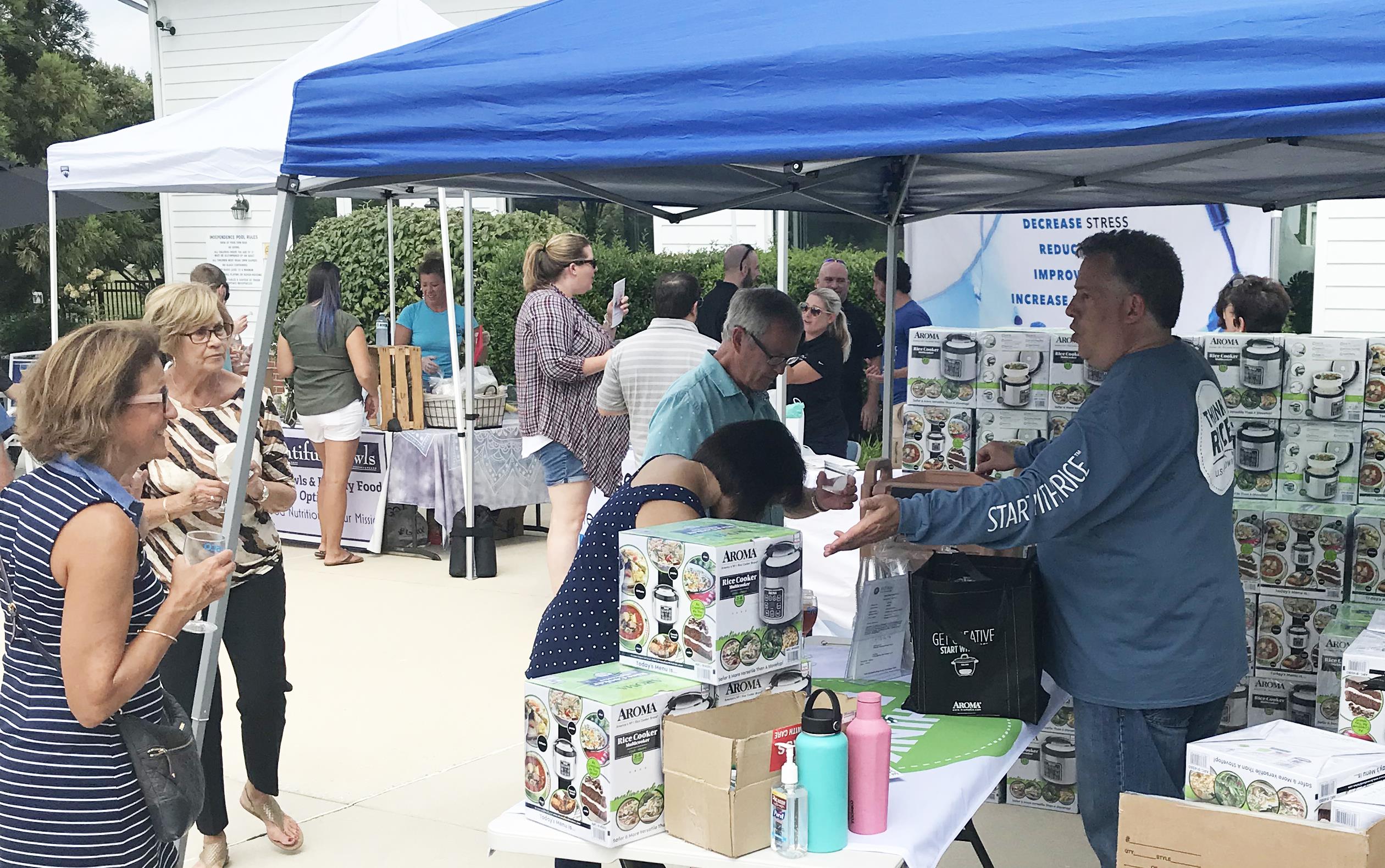 Food Fest booth with blue tent over stacks of rice cookers in boxes and on tables, man in blue shirt gestures to people walking by