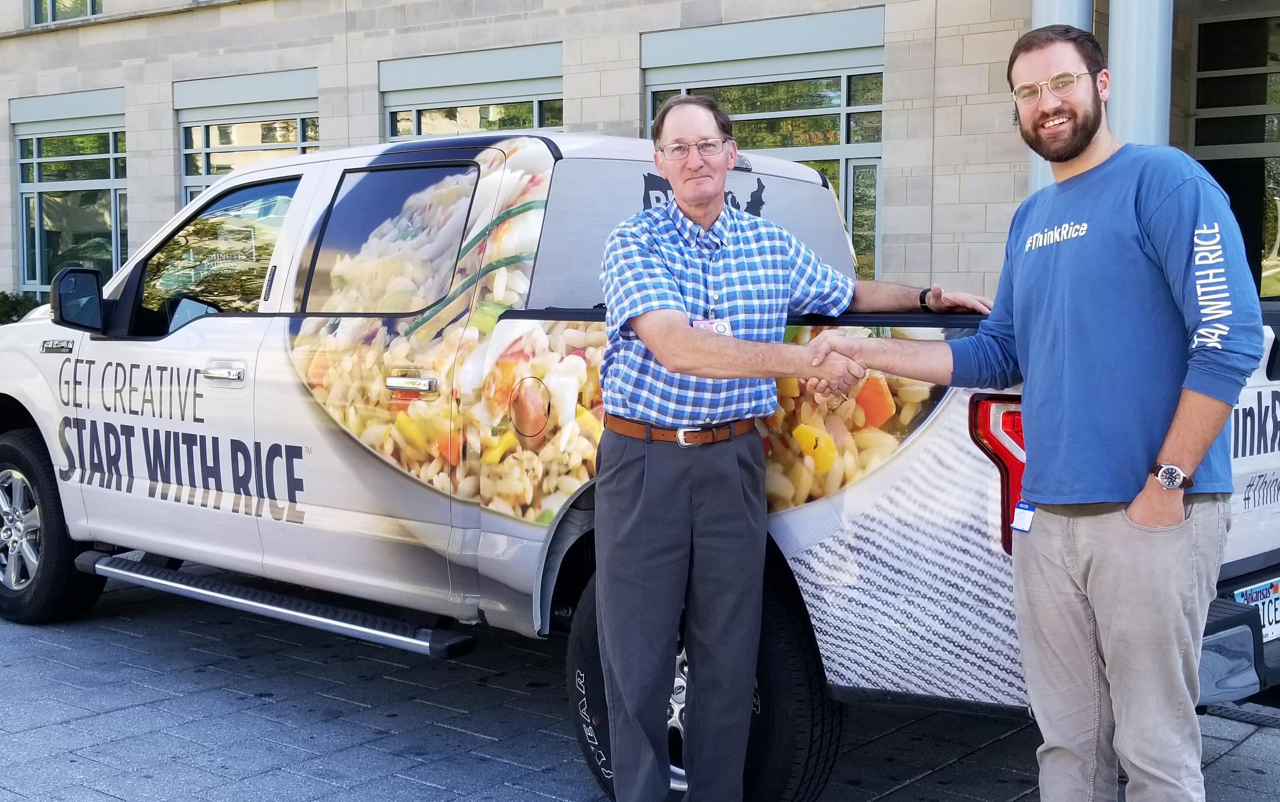 Two men shake hands while standing in front of pickup truck with rice messaging