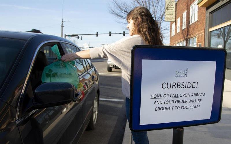 Woman on sidewalk hands bag of food to person in car, signage reads: "Curbside! Honk or call upon arrival & order will be brought to your car!"