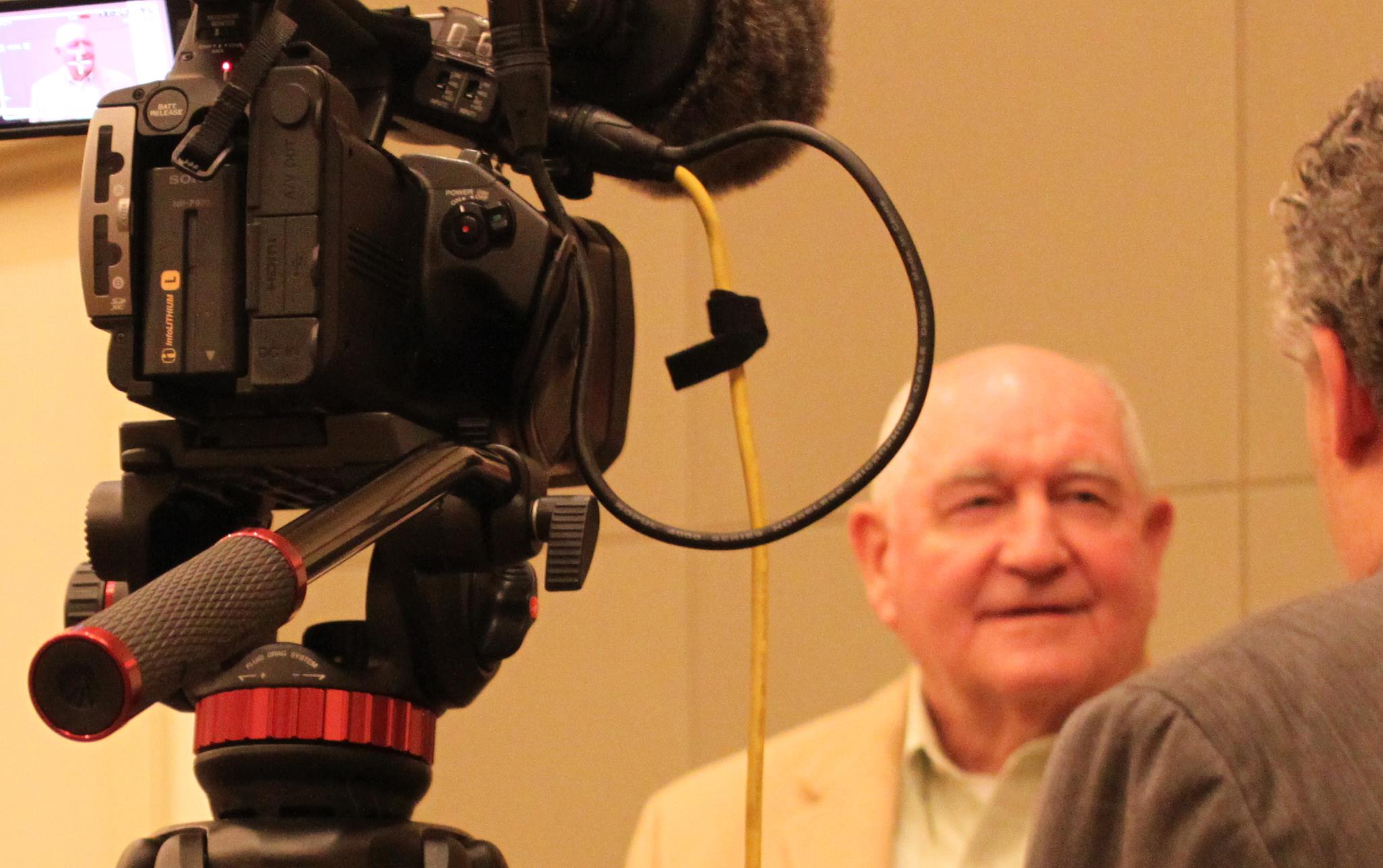 Bald white man stands in front of video camera being interviewed by man wearing a suit