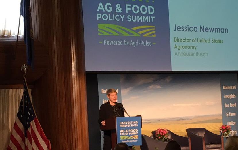 Woman at podium, American flag to the left, under banner text "Harvesting Perspectives Ag & Good Policy Summit"