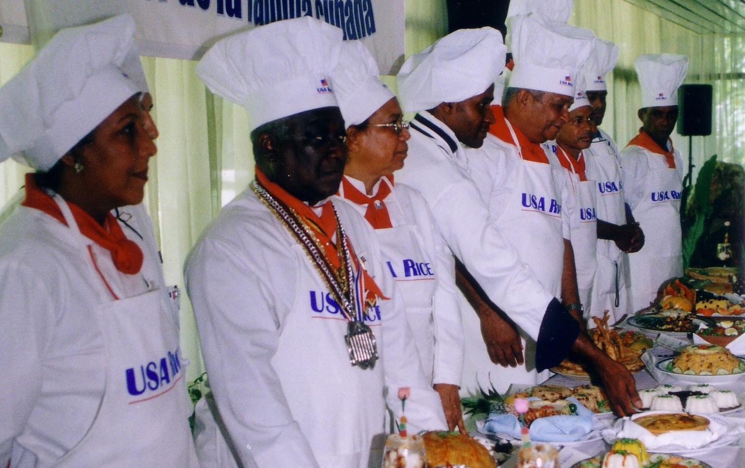 Long line of chefs wearing toques stand behind table laden with food, USA Rice banner hangs above their heads