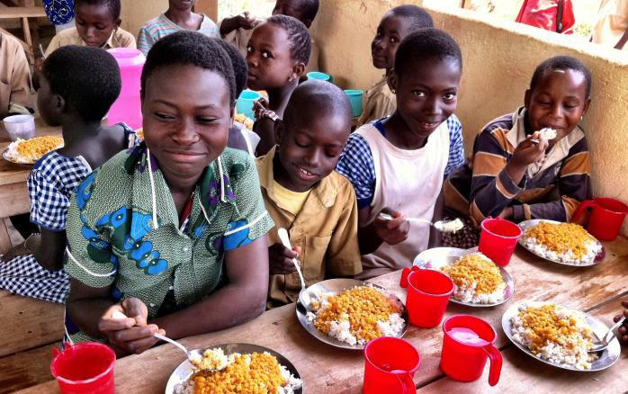 Black children eat lunch of beans and rice at wooden tables 