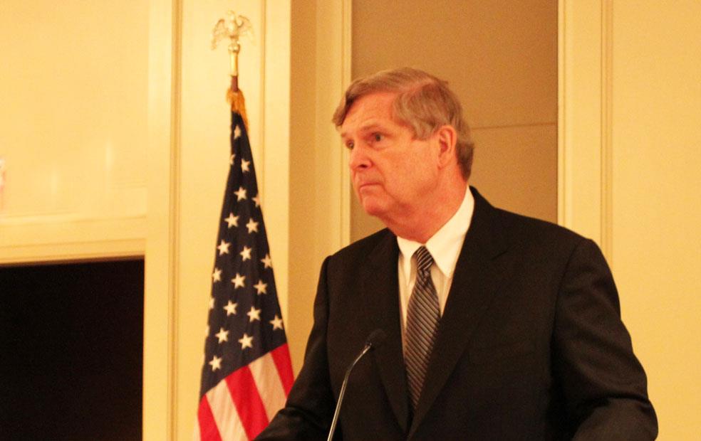 White man in business suit stands in front of  American flag