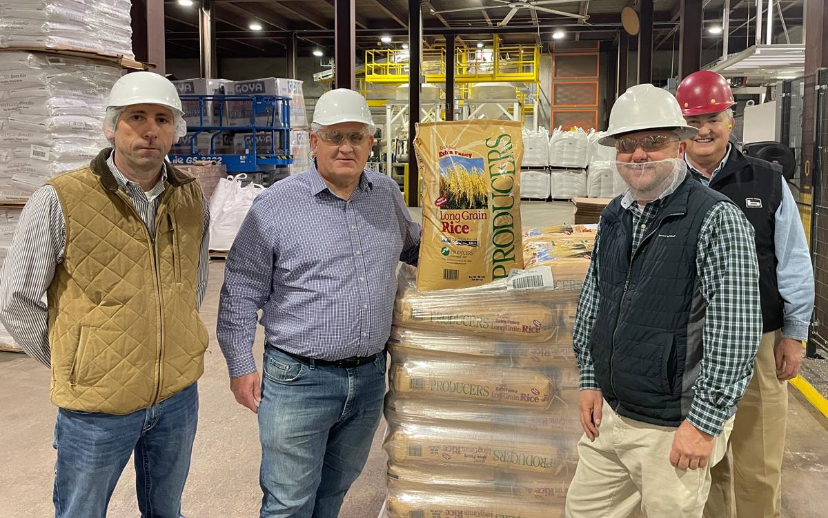 GT-Thompson-Tours-Producers-Rice-Mill, stands with other men wearing hard hats on mill floor surrounded by pallets filled with bags of rice