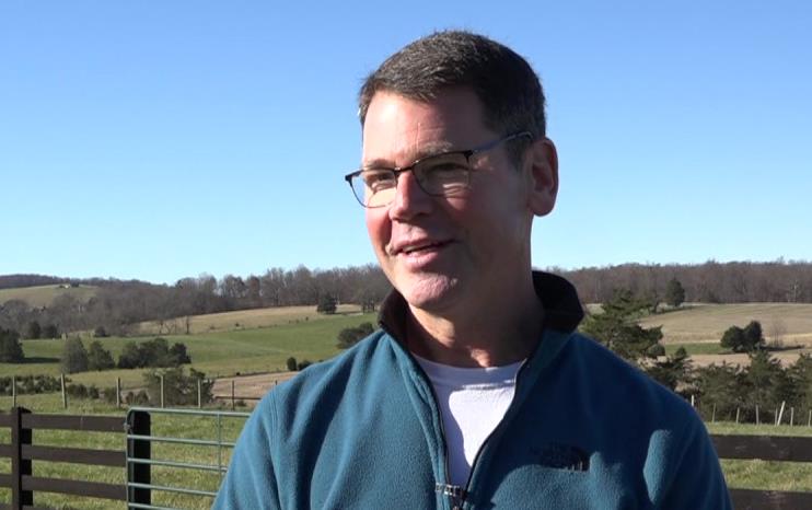 White man wearing glasses & blue sweater standing in fenced in field among rolling hills