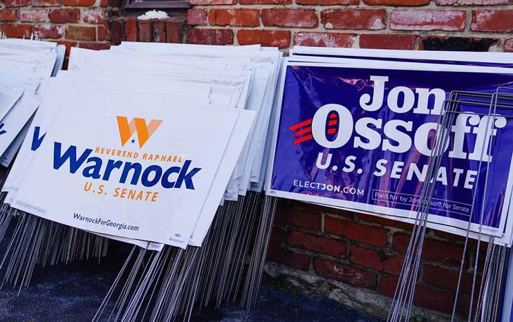 Campaign signs for Raphael Warnock and Jon Ossoff stacked against a brick wall