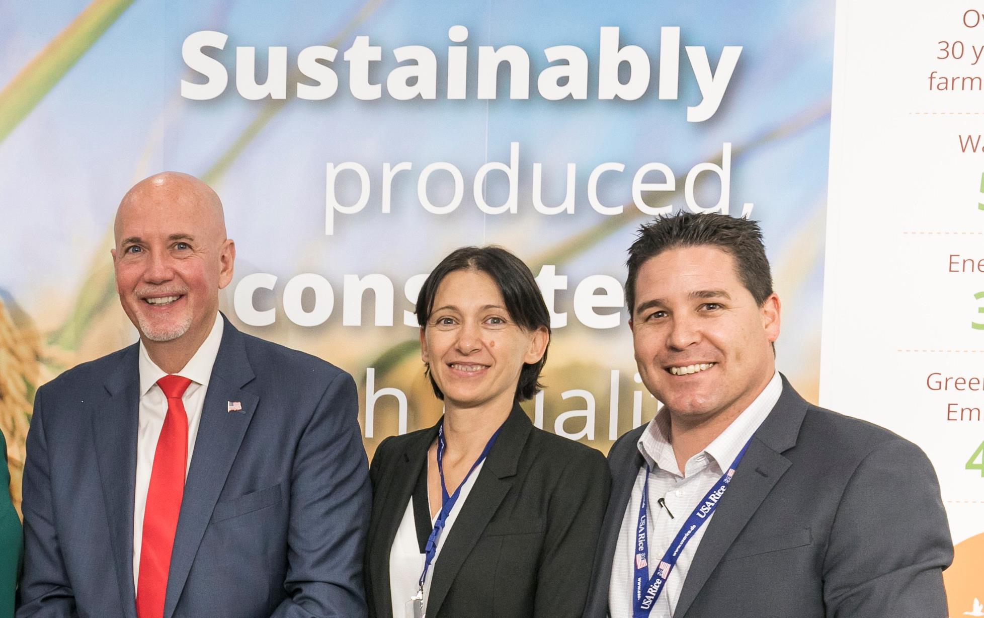 Group shot of people wearing business attire standing in front a poster of rice in the field with sustainability messaging, Kolnmesse photo 