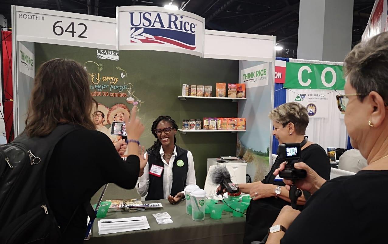 Woman wearing glasses & a big grin, seated at USA Rice booth, being interviewed and filmed by three more woman holding cameras and microphones