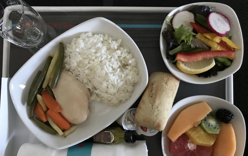 Airline food displayed in small white containers on tray including white rice, colorful fruit and vegetable salads, bread roll, and napkin